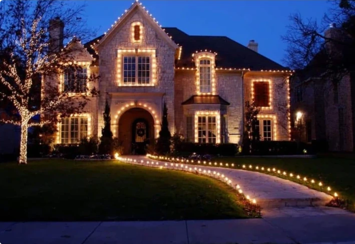 Large brick residence with warm lights along the eaves and a lit driveway